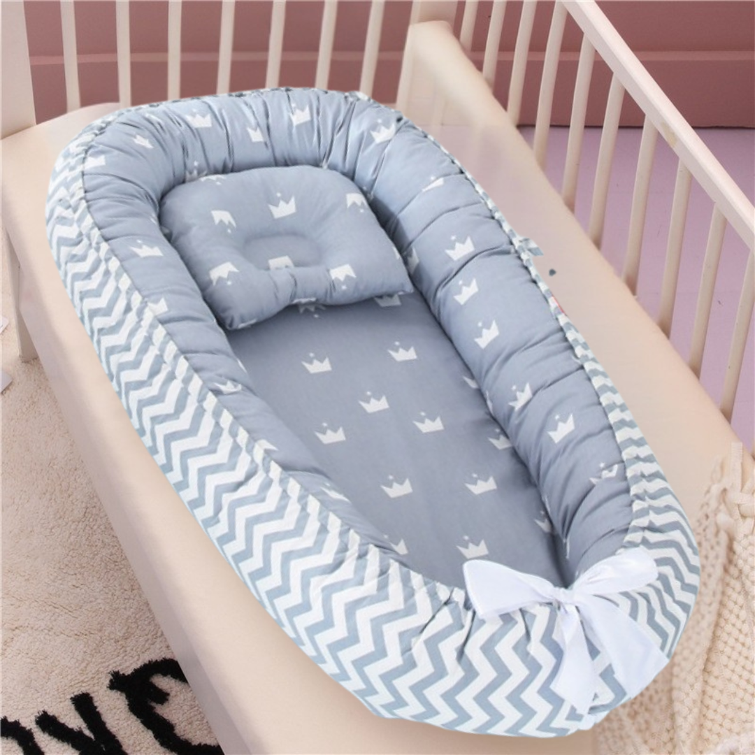 Le réducteur de lit bébé Dodo Cocoon en bleu clair doux avec un motif de couronne et une bordure à chevrons repose dans un berceau pâle, avec un oreiller assorti, un nœud blanc en bas et une partie d'une couverture en tricot crème à proximité.