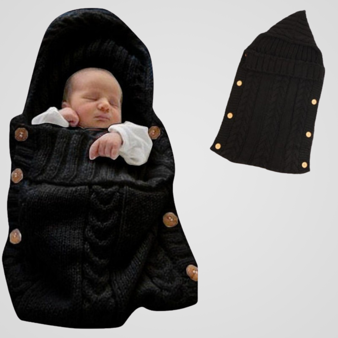 Un bébé endormi est enveloppé dans la Couverture Emmaillotage | Petit Rêve noire en tricot, ornée de boutons en bois. À côté de lui, une couverture à plat met en valeur la forme et le design élégants de ce cocon de douceur.
