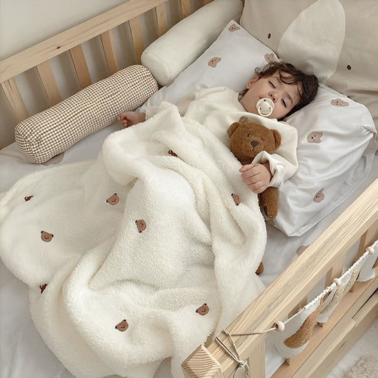 Un tout-petit, muni d'une tétine, est allongé dans un berceau en bois, câlinant un ours en peluche marron, recouvert de la Couverture bébé | P'tit Câlin™, ornée de petits visages d'ours. Le berceau comprend également des coussins moelleux et un oreiller à imprimé animal pour plus de confort.