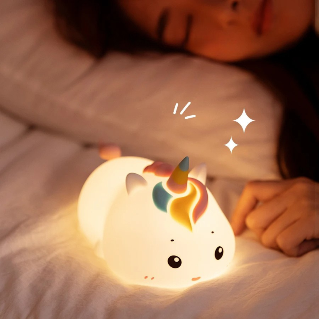 Une personne dort à côté de la veilleuse bébé magique UniGlow™ sur un lit. Cette veilleuse en forme de licorne, avec sa corne colorée, émet une lueur douce et chaleureuse avec des paillettes illustrées, assurant des nuits paisibles.