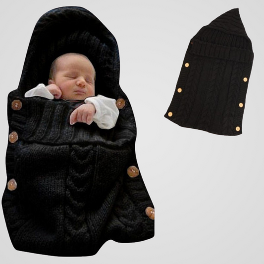 Un bébé endormi est enveloppé dans la Couverture Emmaillotage | Petit Rêve noire en tricot, ornée de boutons en bois. À côté de lui, une couverture à plat met en valeur la forme et le design élégants de ce cocon de douceur.