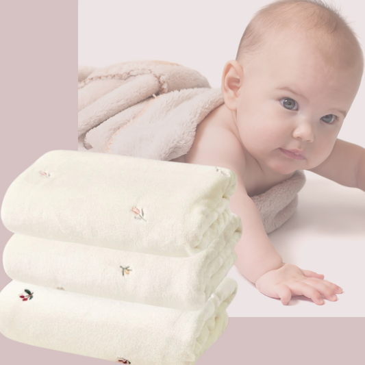 Un bébé est allongé sur une Couverture bébé | Tulipes™ en polaire ultra douce, le regard tourné vers l'avant. Au premier plan, trois serviettes blanches soigneusement pliées, ornées de motifs floraux, sont empilées. L'arrière-plan est rose pâle.