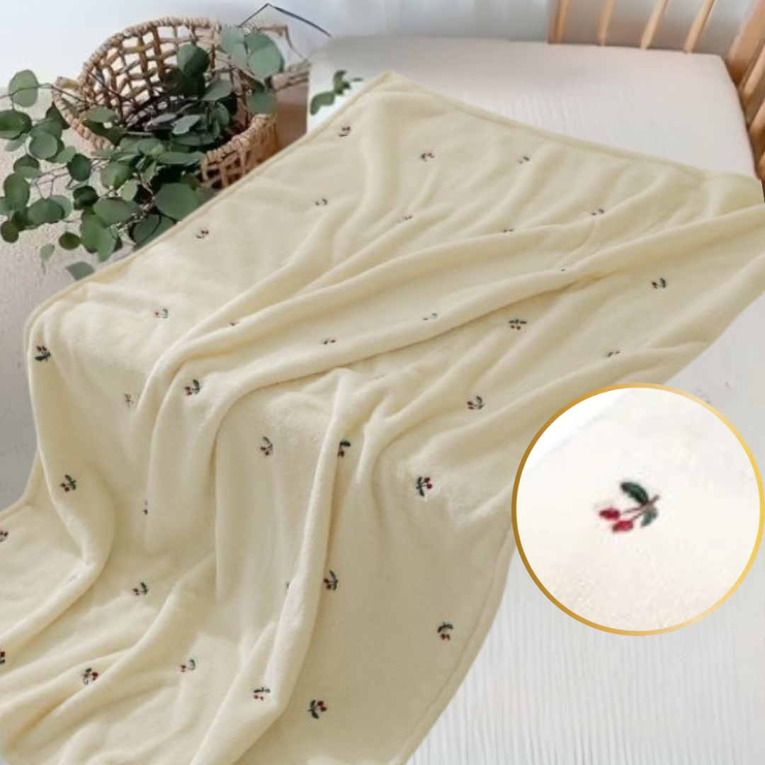 La Couverture bébé | Velours Câlin™ est une couverture bébé en velours ultra-doux jaune clair, ornée de motifs de cerises brodés, posée sur un lit blanc. Un gros plan révèle les cerises rouges et vertes ; un panier en osier garni de verdure trône à proximité.
