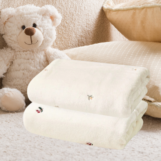 Une Couverture bébé | Velours Câlin™ pliée en velours doux repose sur une surface beige à côté d'un ours en peluche blanc et d'un oreiller crème texturé, créant une ambiance chaleureuse dans la chambre.
