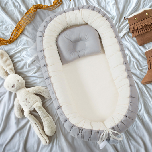 Voici une vue de dessus élégante d'un réducteur de lit pour bébé, doucement niché sur un drap blanc satiné. Un oreiller gris assorti repose à l'intérieur, prêt pour le confort de bébé. À côté, un adorable lapin en peluche crème, des chaussons en daim marron avec des franges, et un ruban doré festif ajoutent une touche chaleureuse et décorative à la scène.