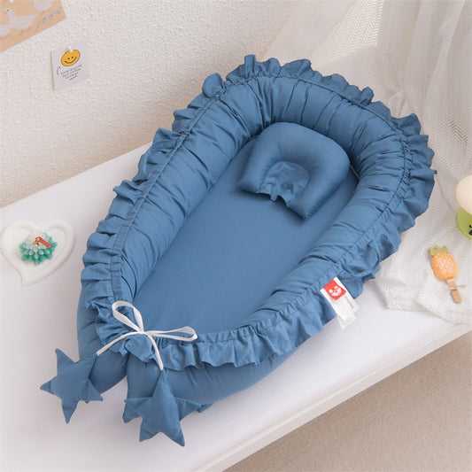 Un adorable cocon de bébé bleu ciel, orné de volants délicats, repose sur un lit blanc. Un oreiller assorti attend la tête de l'enfant. Deux étoiles bleues pendent au pied du nid, attachées par un ruban blanc, ajoutant une touche de fantaisie
