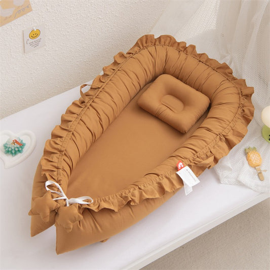Un nid d'ange douillet pour bébé, de couleur camel avec des volants délicats sur les bords. Un petit oreiller rectangulaire assorti repose à l'intérieur, invitant au repos. Le nid est placé sur un fond blanc, suggérant un environnement sûr et confortable pour le nourrisson.