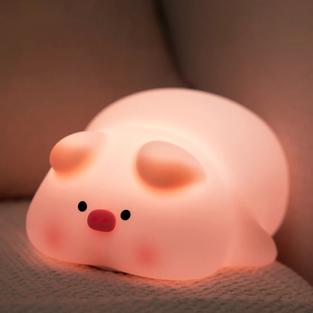 La veilleuse bébé Porcin™, au corps rond, aux joues rougissantes, aux petits yeux noirs, au museau rose et aux oreilles arrondies, émet une douce lueur sur une surface coussinée pour créer une ambiance mignonne et réconfortante.