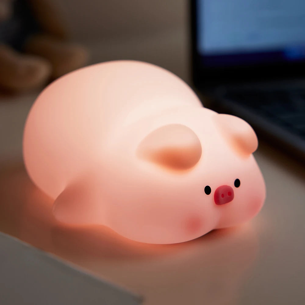 La Veilleuse bébé | Porcin™ est une lampe en forme de cochon aux formes arrondies et à la lumière douce, posée sur un bureau. Sa teinte rose et son expression mignonne offrent une ambiance apaisante. En arrière-plan, un ordinateur portable flou est visible.
