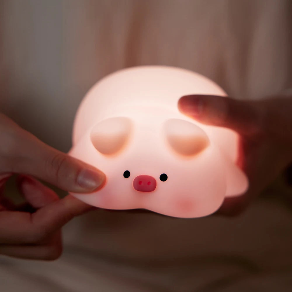 Dans une pièce faiblement éclairée, une personne berce l'adorable veilleuse Porcin™. Cette veilleuse bébé est dotée de petites oreilles et d'un museau rose, se fondant harmonieusement tout en projetant sa douce lumière chaude dans tout l'espace avec son charmant design en forme de cochon.