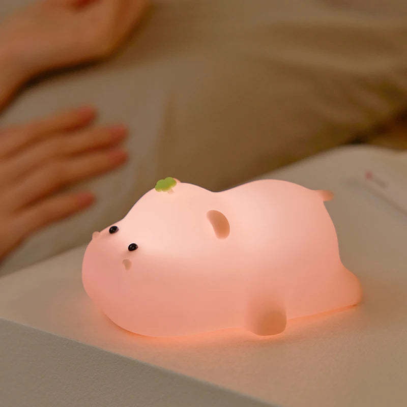 La Veilleuse bébé | Porcin™ est une adorable veilleuse en forme d'animal potelé avec de petites oreilles et une petite feuille sur la tête, projetant une douce lueur rose pêche lorsqu'elle est posée sur une table de chevet, avec une partie du bras d'une personne visible en arrière-plan.