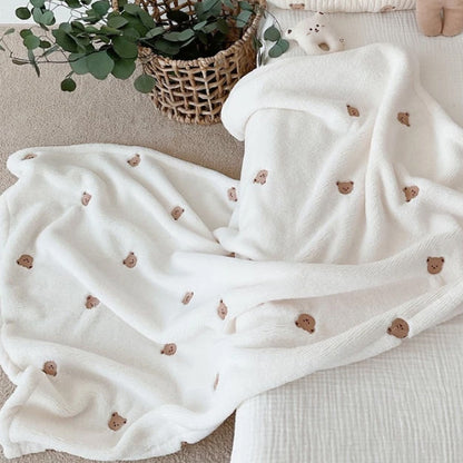 La Couverture bébé | P'tit Câlin™ d'un blanc doux, ornée de petits visages d'ours bruns, est drapée sur un lit à côté d'un panier tressé avec des plantes vertes feuillues sur un sol recouvert de moquette beige.