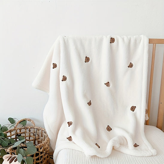 La Couverture bébé | P'tit Câlin™, une douce couverture blanche ornée de petites têtes d'ours bruns, orne un berceau en bois. À proximité, un panier tressé garni de verdure complète le confort douillet et multifonctionnel de cet accessoire indispensable.