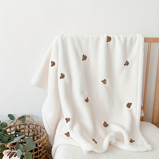 La Couverture bébé | P'tit Câlin™, une douce couverture blanche ornée de petites têtes d'ours bruns, orne un berceau en bois. À proximité, un panier tressé garni de verdure complète le confort douillet et multifonctionnel de cet accessoire indispensable.