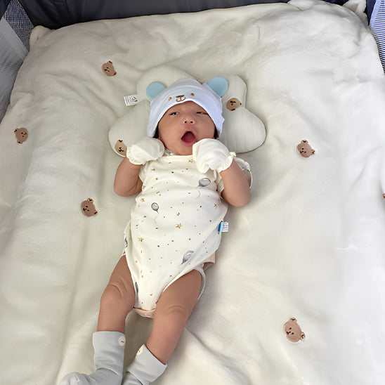 Un bébé en body blanc et bonnet d’ours est allongé sur la couverture bébé P’tit Câlin™, douce et ornée de petits ours, avec un coussin assorti. Ce produit multifonction assure un confort optimal pendant que bébé découvre le monde.