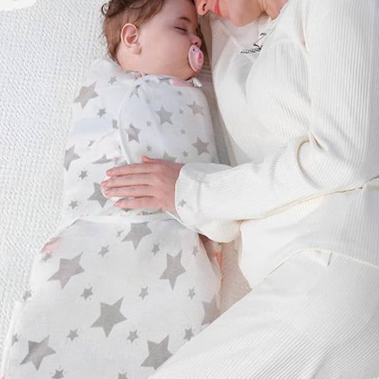 Un bébé enveloppé dans une Couverture emmaillotage bébé | Douce Câline™ à étoiles grises dort paisiblement aux côtés d'un adulte en blanc, tous deux sur une surface blanche texturée. Le bébé porte une tétine rose.