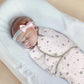 Un nouveau-né endormi enveloppé dans une Couverture emmaillotage bébé Douce Câline™ avec des étoiles et des lunes grises est allongé sur une surface rembourrée blanche, profitant d'un sommeil paisible et portant un bandeau léger avec un nœud rose.