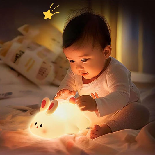 Un bébé est assis sur un lit, jouant intensément avec un BunnyGlow™ de Petitsbeautes. La veilleuse lumineuse en forme de lapin projette une ambiance douce autour de la pièce faiblement éclairée, créant une atmosphère chaleureuse et douillette, avec une petite étoile dessinée visible au-dessus de la tête du bébé, ce qui en fait le compagnon de nuit idéal.