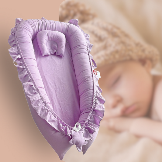 Un confortable nid d'ange lavande, orné de délicats volants, est mis en avant sur un fond doux et rêveur. Un petit oreiller assorti repose à l'intérieur du couffin. En arrière-plan flou, un bébé paisiblement endormi, coiffé d'un bonnet tricoté, suggère le confort et la sécurité de ce lit douillet. La scène évoque la douceur et le bien-être des tout-petits.